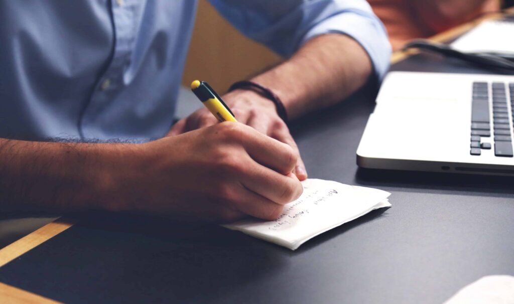 Person writing notes on a notepad beside an open laptop at a desk