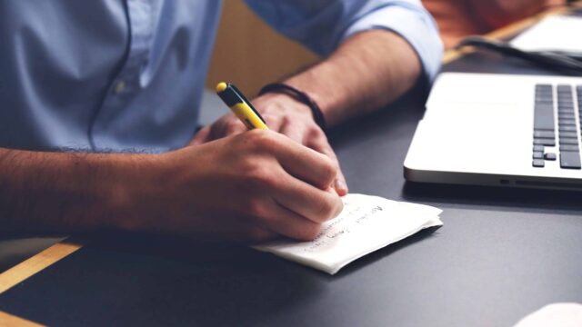 Person writing notes on a notepad beside an open laptop at a desk