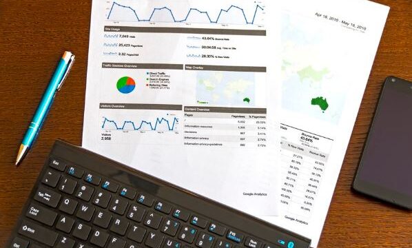 Printed analytics dashboard reports on a desk with keyboard and phone, representing Facebook Ads and AdWords ROI tracking