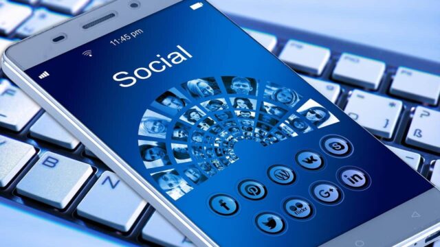 Smartphone resting on a keyboard displaying a social media app with platform icons including Twitter and LinkedIn