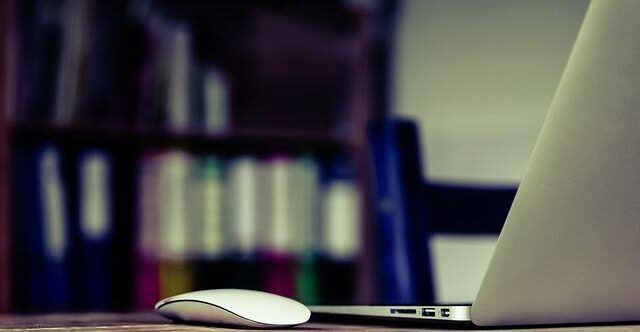 Laptop and wireless mouse on a desk with colourful binder folders in the background