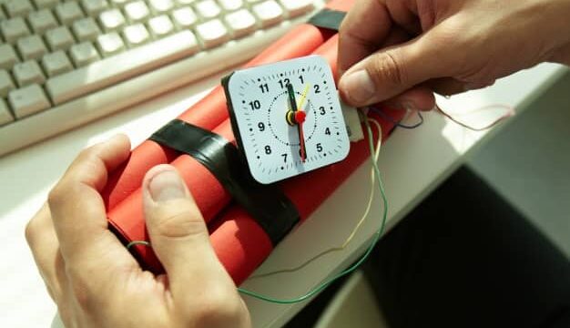Hands holding a red dynamite bundle with a clock timer next to a computer keyboard