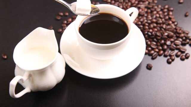 Cup of black coffee with sugar cube on spoon, milk jug and scattered coffee beans on dark surface