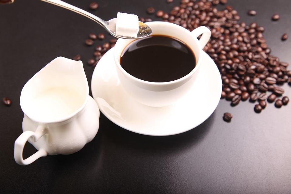 Cup of black coffee with sugar cube on spoon, milk jug and scattered coffee beans on dark surface