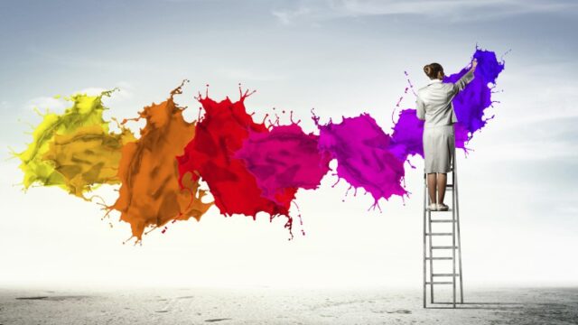 Woman on a ladder painting a vivid multicolour paint splash across a grey sky, representing creative work