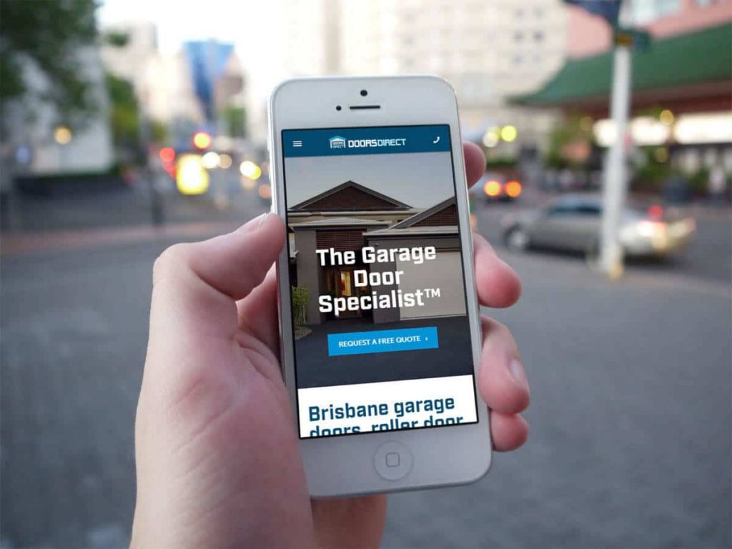Hand holding a smartphone displaying the Doors Direct mobile website, with a Brisbane street scene in the background