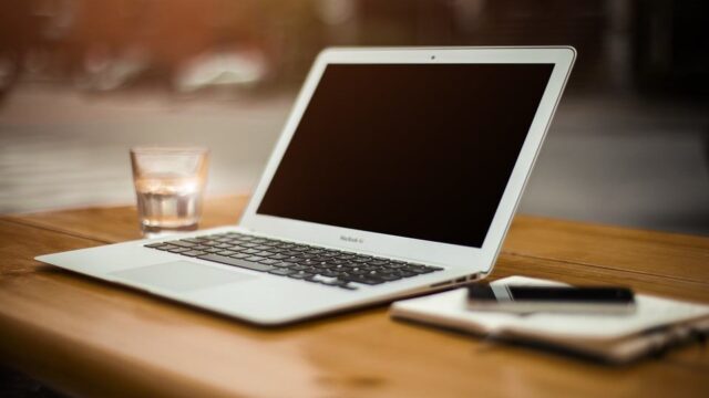 MacBook Air open on a wooden table beside a glass of water and a notebook, representing blogger outreach tools