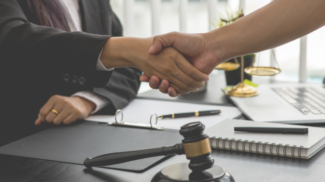 Two professionals shaking hands at a desk with gavel and scales of justice, representing legal client relationships