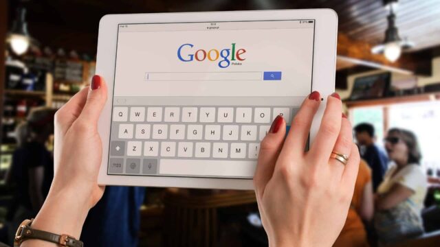 Woman using both hands to type on an iPad showing Google search homepage with on-screen keyboard