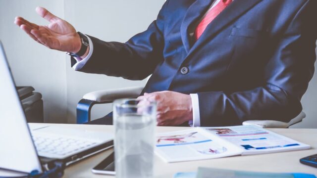 Business professional in a suit gesturing at a desk with reports and a laptop, illustrating thought leadership in your industry