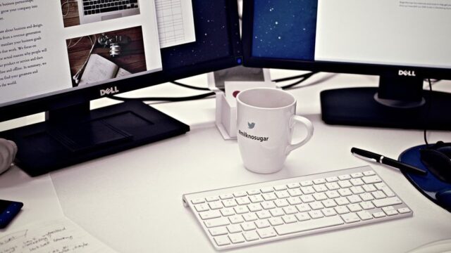 Designer's dual-monitor desk setup with website mockups on screen, keyboard and Twitter mug in foreground