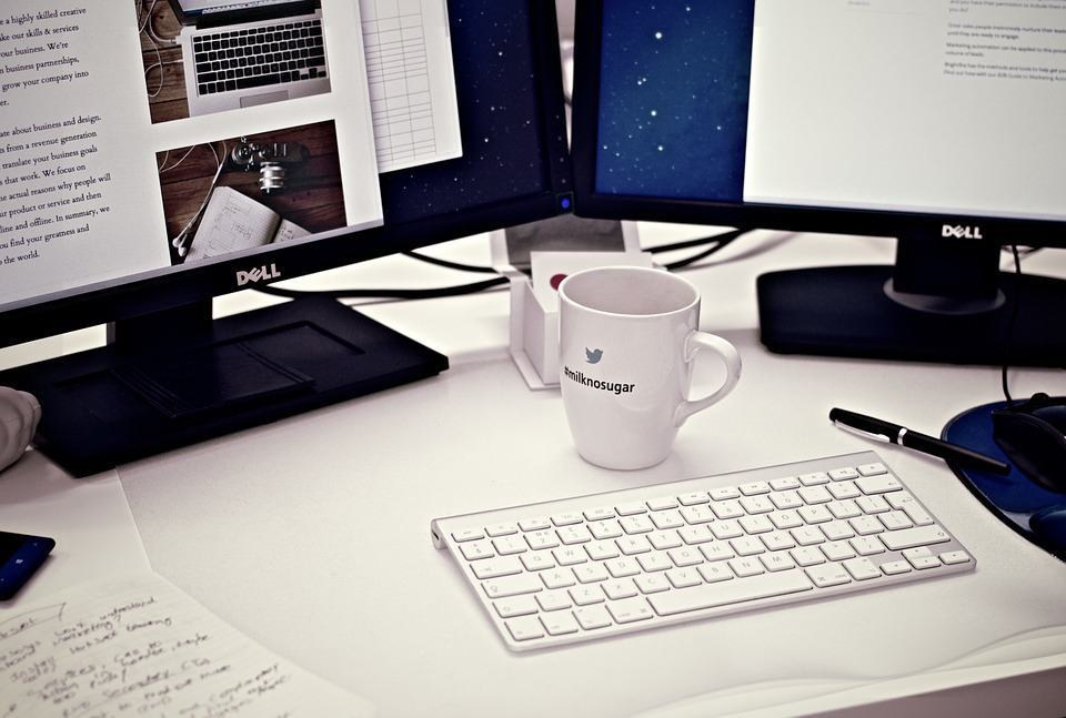Designer's dual-monitor desk setup with website mockups on screen, keyboard and Twitter mug in foreground