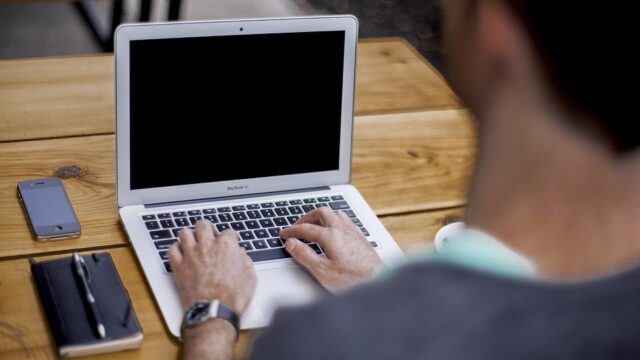 Man typing on a MacBook Air at a wooden desk with a notepad and smartphone nearby, viewed from behind