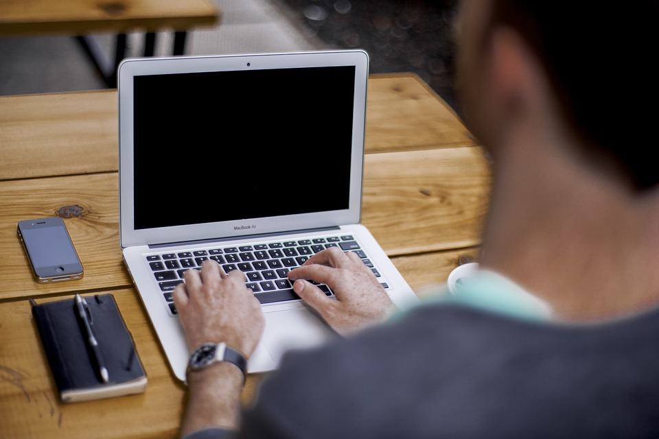 Man typing on a MacBook Air at a wooden desk with a notepad and smartphone nearby, viewed from behind