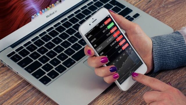 Woman holding a smartphone displaying a stock market app in front of an open MacBook Air