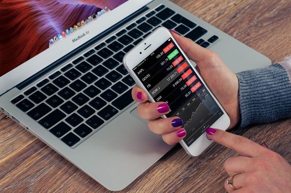 Woman holding a smartphone displaying a stock market app in front of an open MacBook Air