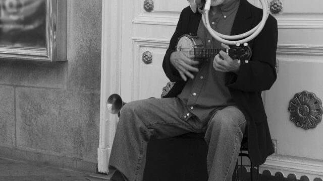 Street musician sitting outside a building playing a ukulele with a horn attached, illustrating a one-man-band