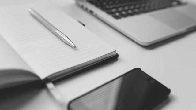 Black and white flat-lay of a notebook with a pen, laptop, and smartphone on a white desk