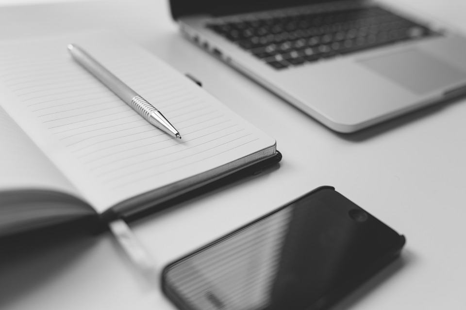 Black and white flat-lay of a notebook with a pen, laptop, and smartphone on a white desk
