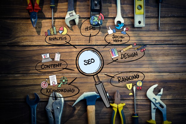 Magnifying glass labelled SEO on a wooden workbench with speech bubbles showing SEO components: content, keywords, ranking, design, HTML, and social media
