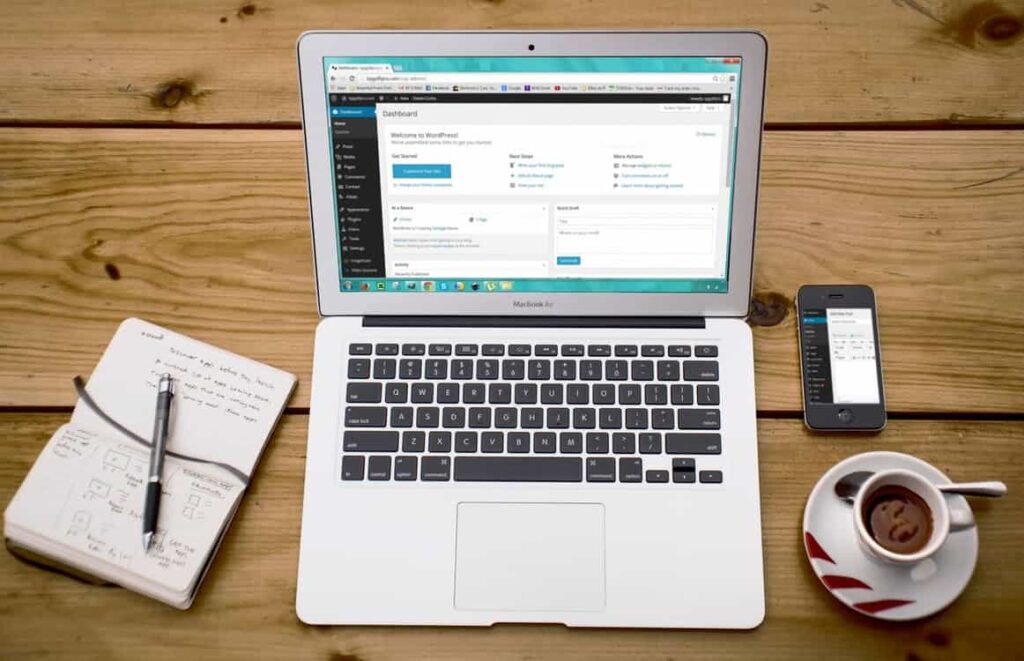MacBook Air showing the WordPress dashboard on a wooden desk with a coffee cup and notebook