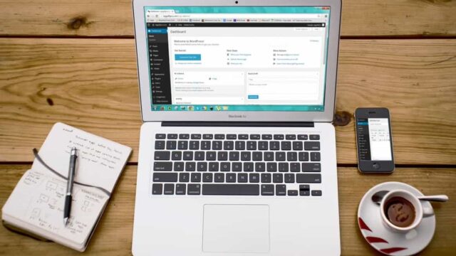 MacBook Air showing the WordPress dashboard on a wooden desk with a coffee cup and notebook