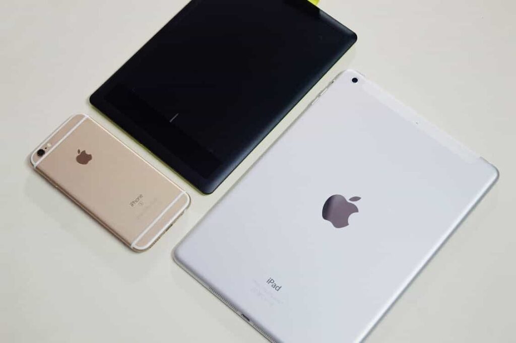 iPhone, black tablet and white iPad laid face-down on a white surface showing Apple device range