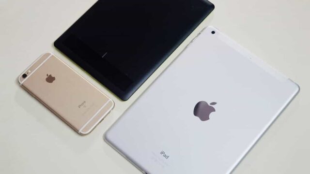 iPhone, black tablet and white iPad laid face-down on a white surface showing Apple device range