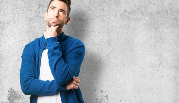 Young man in a blue jacket looking upward with hand on chin in a thoughtful pose