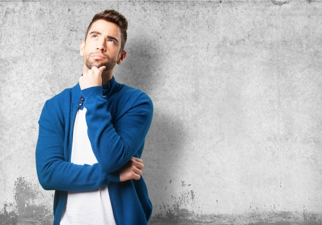Young man in a blue jacket looking upward with hand on chin in a thoughtful pose