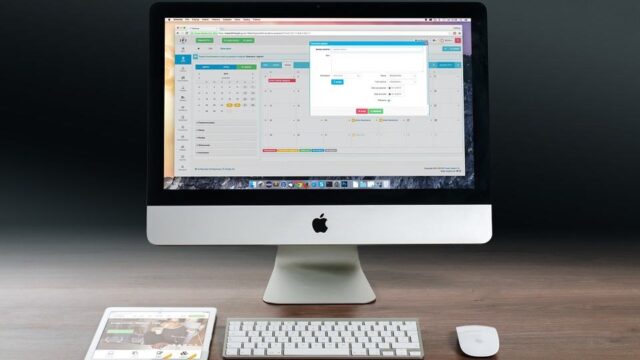 iMac desktop showing a web application calendar dashboard with a tablet and keyboard on a desk